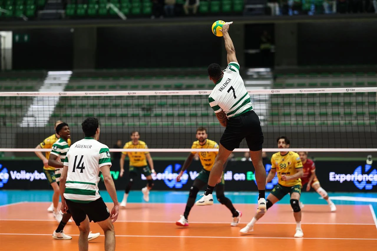 Kadr z meczu Sporting - Aluron CMC Warta Zawiercie Zawodnik drużyny w zielono-białych pasach unosi się w powietrzu wykonując atak podczas meczu siatkówki na hali, naprzeciw widoczna drużyna ubrana na żółto, skoncentrowana na obronie.