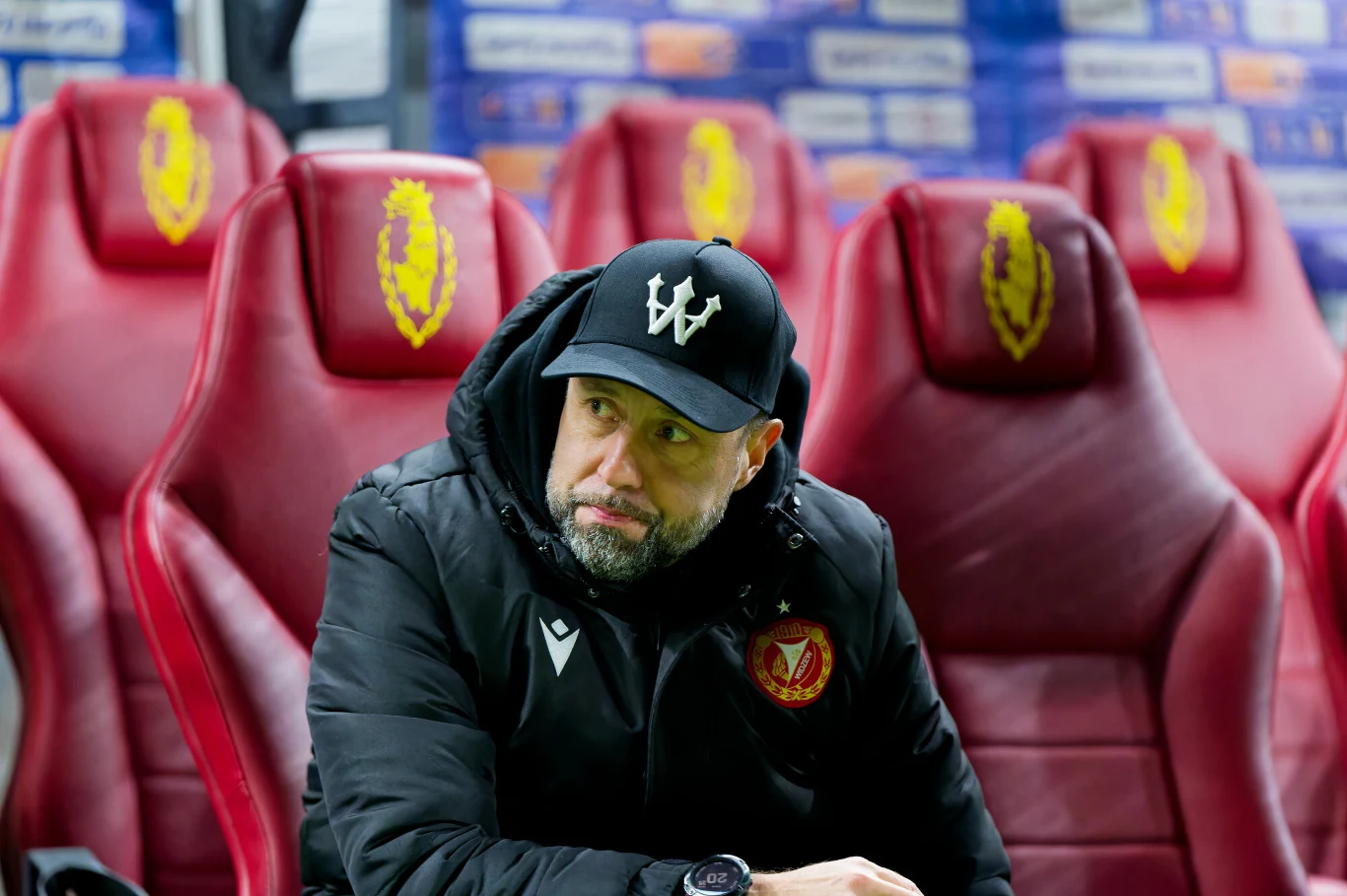 Trener piłkarski w czarnej czapce z daszkiem oraz kurtce z herbem klubu siedzi samotnie na czerwonym fotelu na stadionie, za nim rząd pustych siedzeń i nieostre tło z elementami stadionowymi.