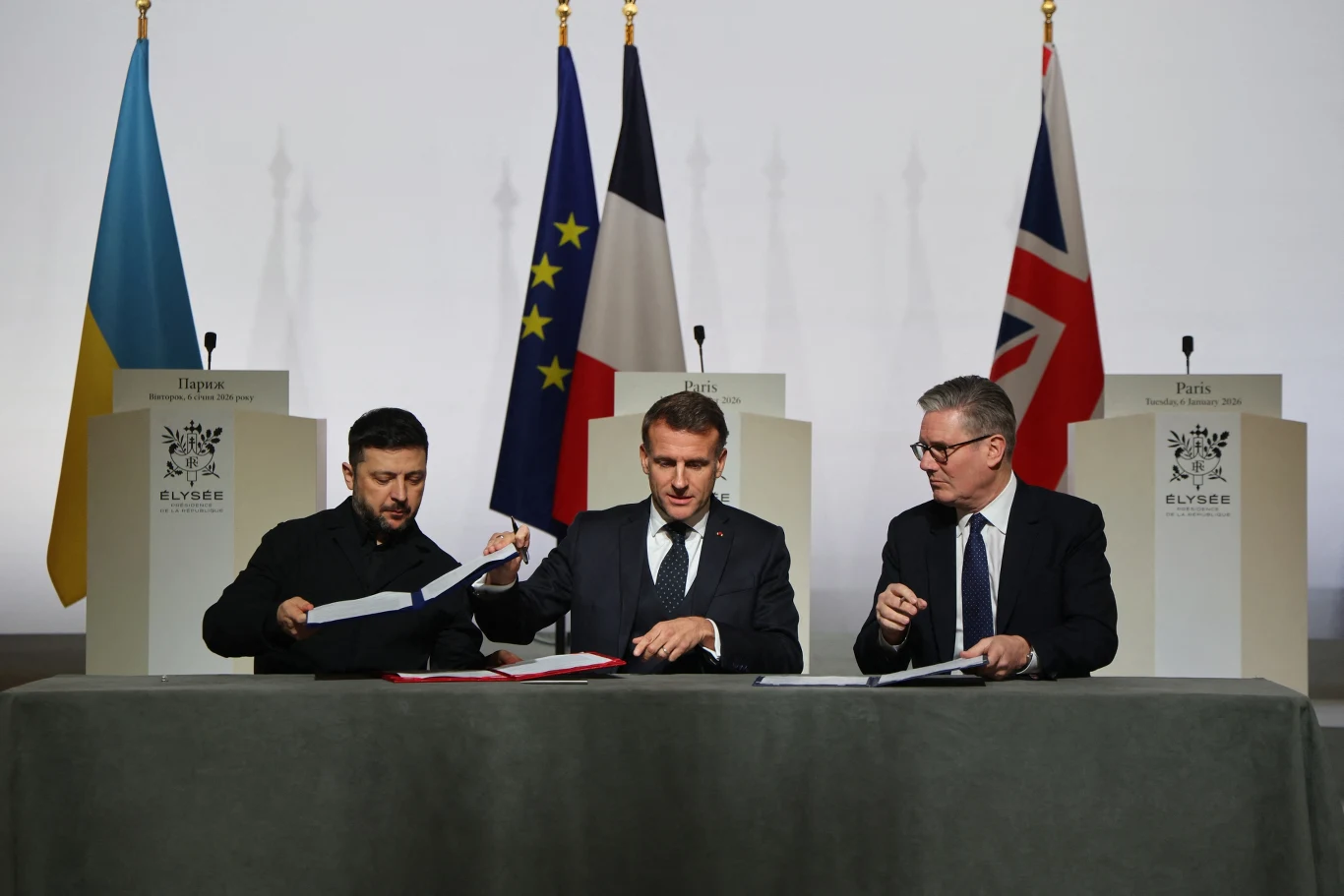 Trzech mężczyzn w garniturach siedzi przy stole i podpisuje dokumenty na tle flag Ukrainy, Unii Europejskiej, Francji oraz Wielkiej Brytanii. Na stole widoczne są teczki i mikrofony, a w tle znajdują się mównice z herbami.