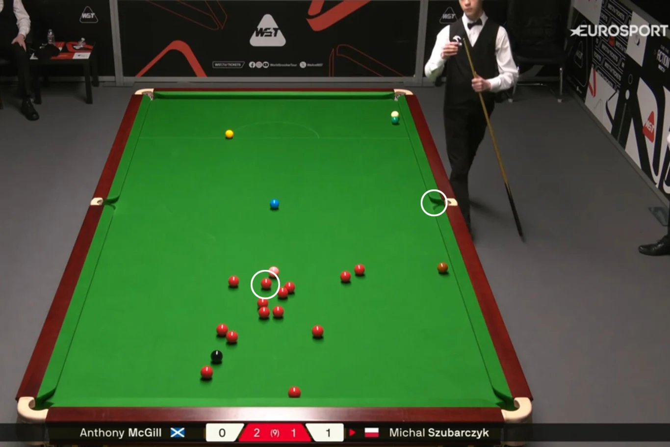 Stół bilardowy z rozmieszczonymi bilami snookerowymi, dwóch zawodników w eleganckich strojach, jeden z nich przygotowuje się do uderzenia kijem, na ekranie wyświetlane są nazwiska zawodników i wynik meczu.