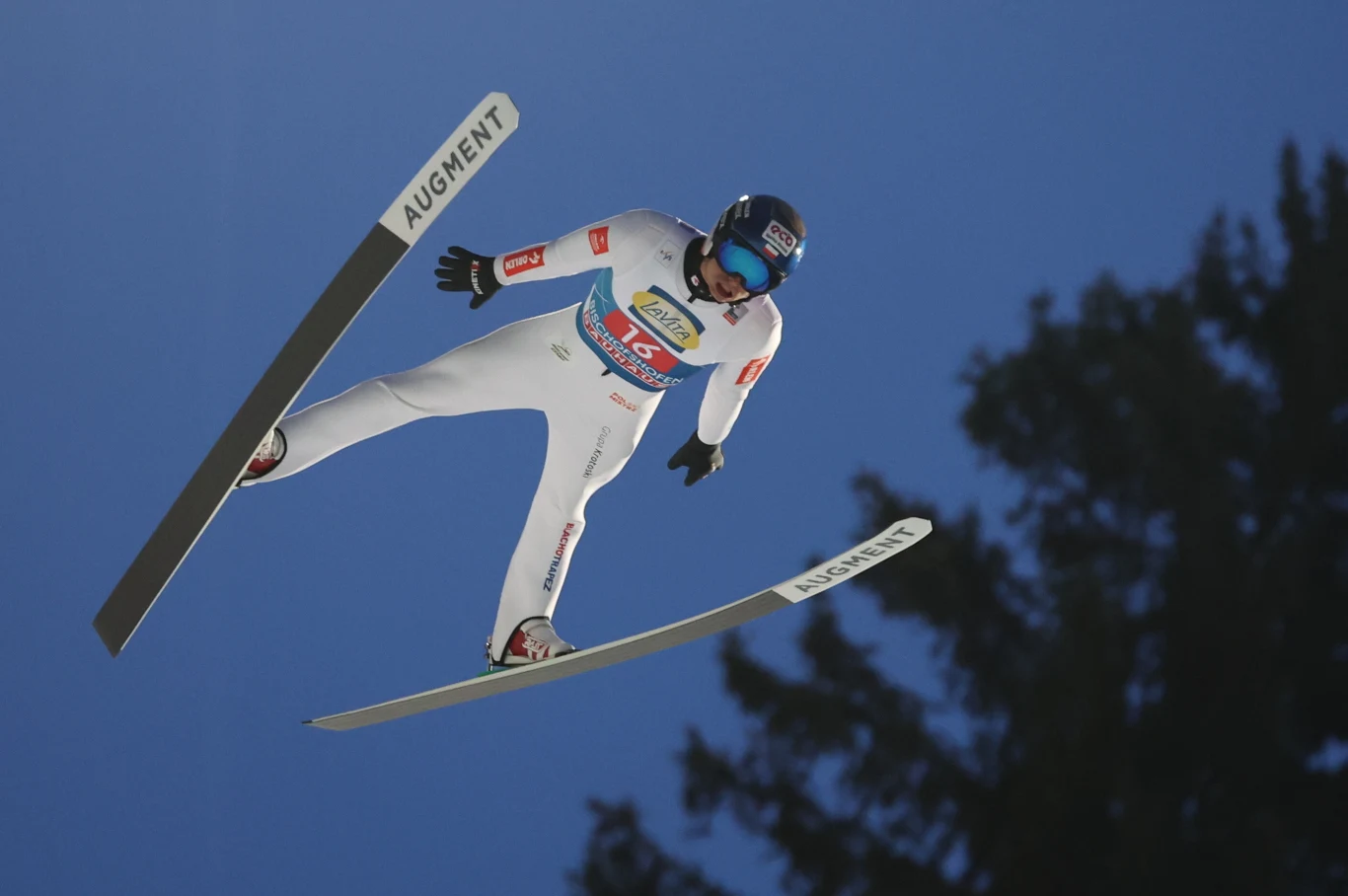 Skoczek narciarski w locie na tle nieba, ujęty w białym kombinezonie z widocznymi logotypami sponsorów, nad ciemnymi zarysami drzew.