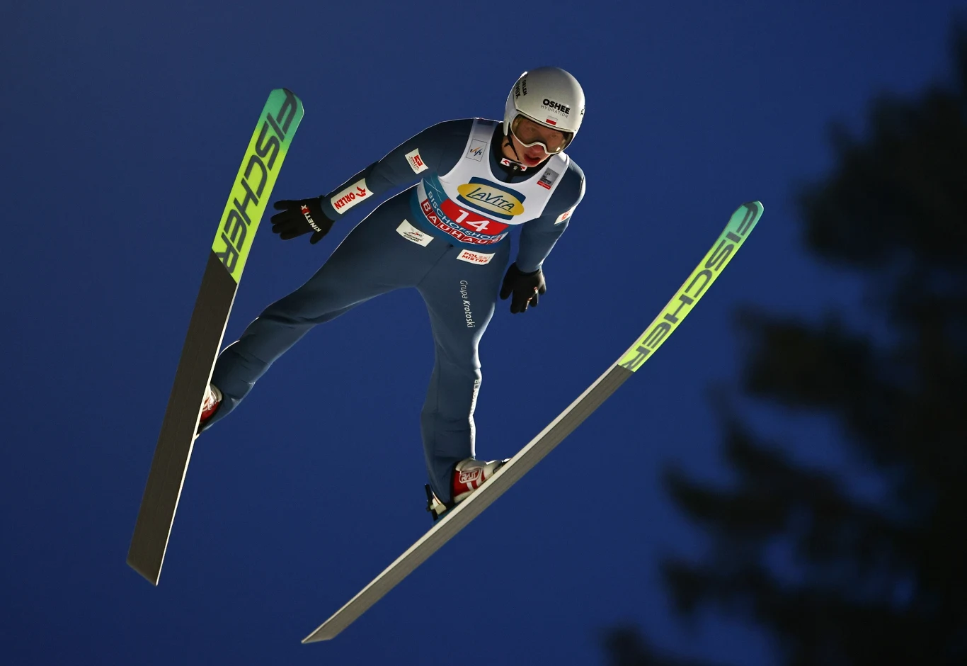 Skoczek narciarski w locie na tle granatowego nieba, ubrany w profesjonalny kombinezon z widocznymi oznaczeniami sponsorów, w kasku i goglach, narty rozstawione szeroko w charakterystycznej pozycji V.