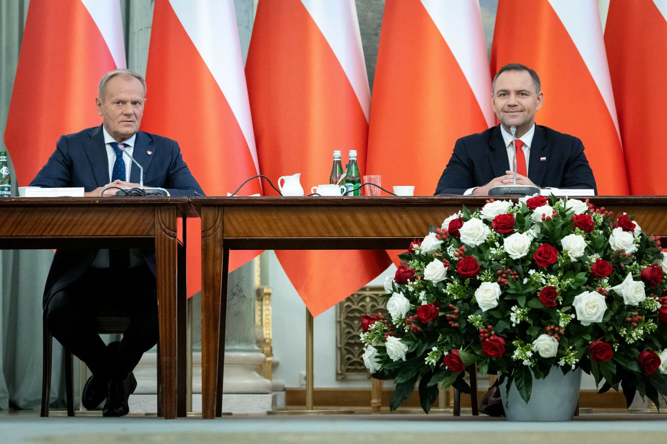 Dwóch mężczyzn siedzących za dużymi stołami ozdobionymi biało-czerwonymi kwiatami, tło stanowią ustawione gęsto polskie flagi.