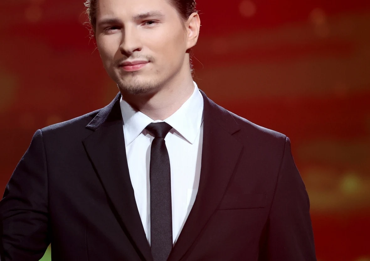 Adam Stachowiak rozstał się z żoną Młody mężczyzna w eleganckim, czarnym garniturze z białą koszulą i czarnym krawatem, stoi na tle czerwono-pomarańczowego, lekko rozmytego tła.