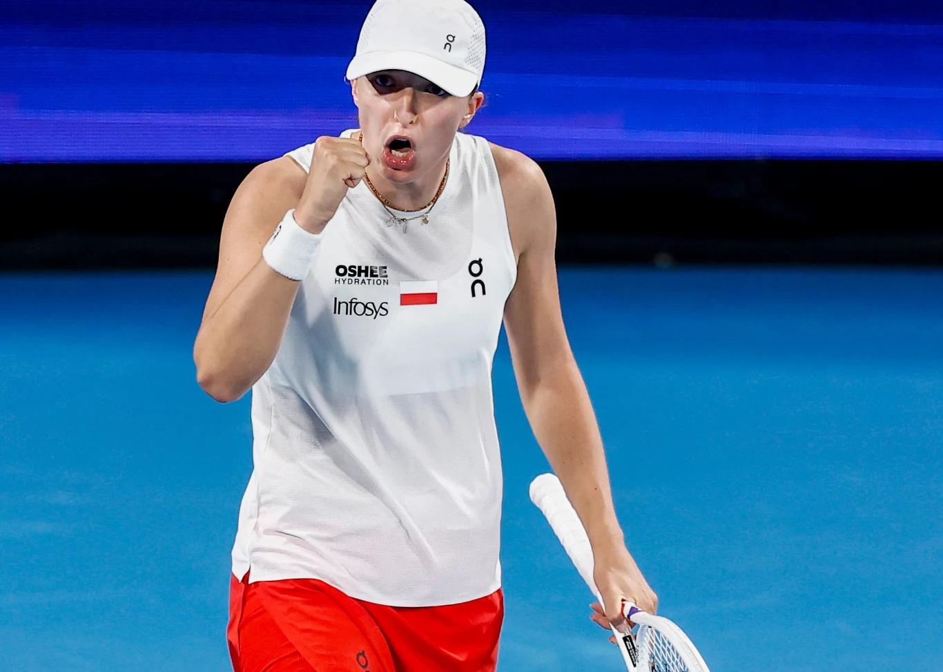 Iga Świątek podczas United CUp 2025 Tenisistka w białej koszulce i czerwonych spodenkach świętuje punkt na niebieskim korcie, trzymając rakietę tenisową i zaciskając pięść z wyrazem determinacji na twarzy.