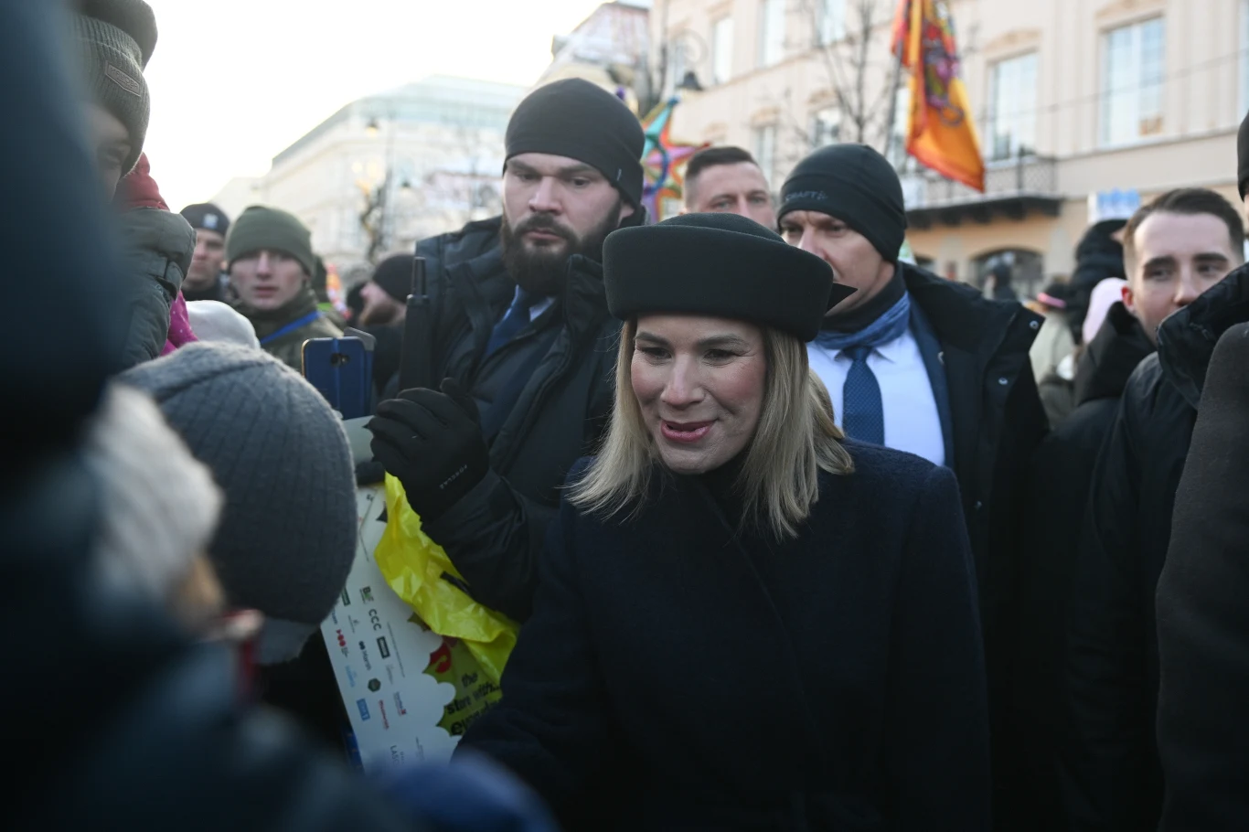 Uśmiechnięta kobieta w czarnym berecie otoczona przez grupę ludzi w zimowych ubraniach podczas zgromadzenia na świeżym powietrzu w mieście, w tle widoczne flagi i budynki.