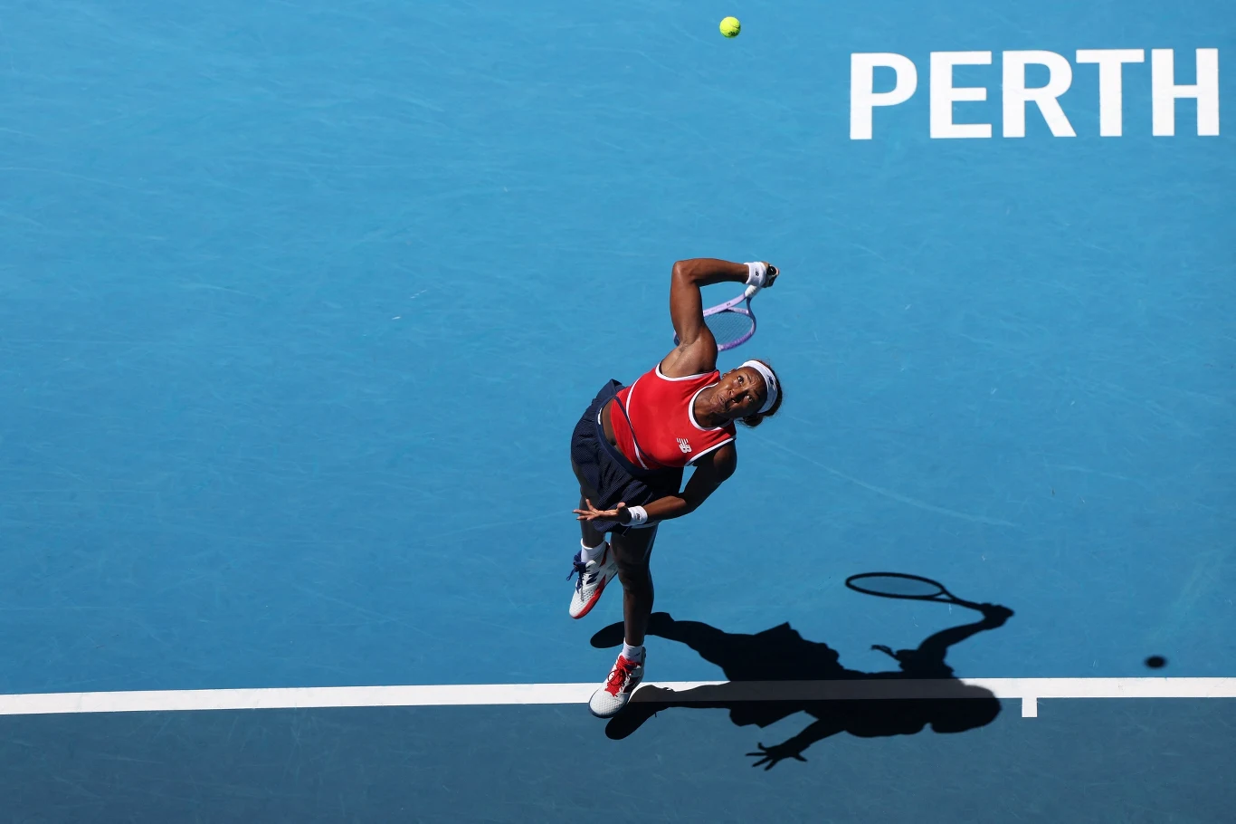 Zawodniczka podczas serwisu na kortach tenisowych, ujęcie z góry, w tle niebieska nawierzchnia kortu oraz napis PERTH w prawym górnym rogu.