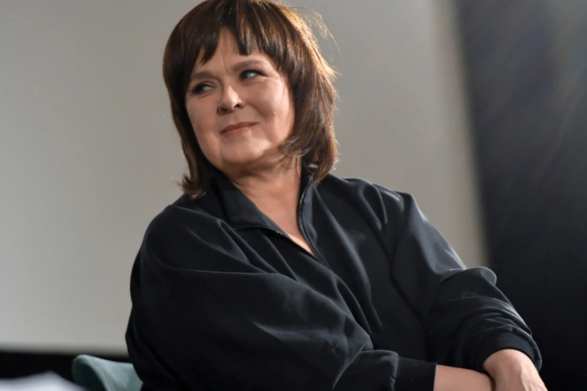 Edyta Jungowska Kobieta z krótkimi brązowymi włosami ubrana w ciemną kurtkę siedząca bokiem na tle rozmytego, neutralnego tła z delikatnym uśmiechem na twarzy.