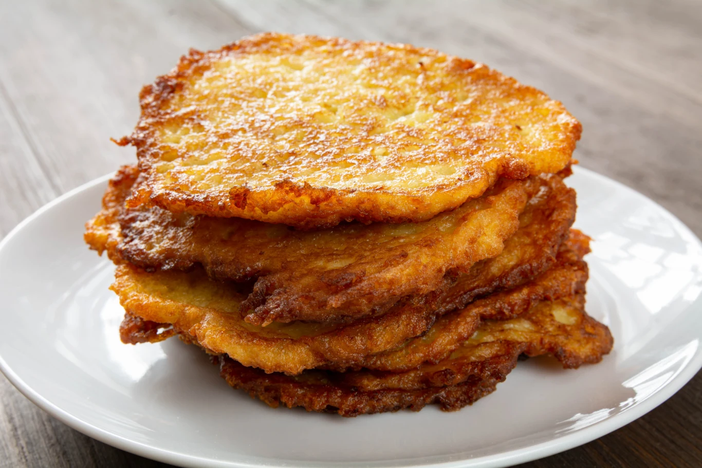 Stos złocistych, chrupiących placków ziemniaczanych ułożonych na białym talerzu na drewnianym stole.