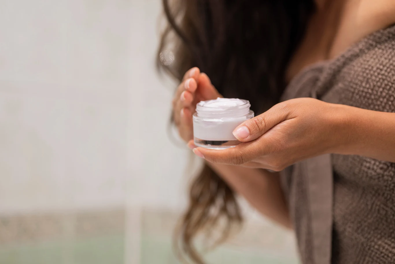 Kobieta trzyma w dłoniach otwarte opakowanie kremu, owinięta w ręcznik, z długimi, ciemnymi włosami. W tle rozmyte kafelki łazienkowe, akcent na pielęgnację skóry i chwilę relaksu.