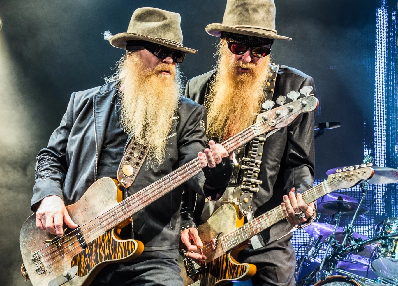 Dusty Hill i Billy Gibbons z ZZ Top Dwóch mężczyzn z długimi brodami i w ciemnych okularach, ubranych w czarne stroje i kapelusze, grających na gitarach elektrycznych na tle kolorowego oświetlenia scenicznego.