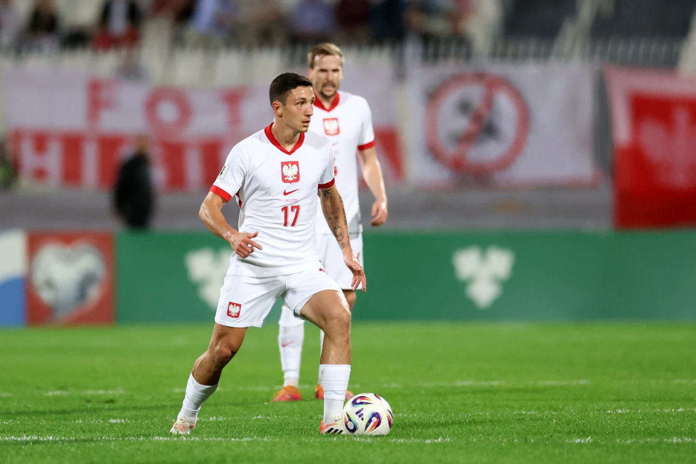 Bartosz Slisz w barwach reprezentacji Polski Piłkarz reprezentacji Polski w białym stroju z numerem 17 prowadzi piłkę podczas meczu piłkarskiego na zielonej murawie. W tle widoczny inny zawodnik w podobnym stroju oraz fragment trybun i transparenty.