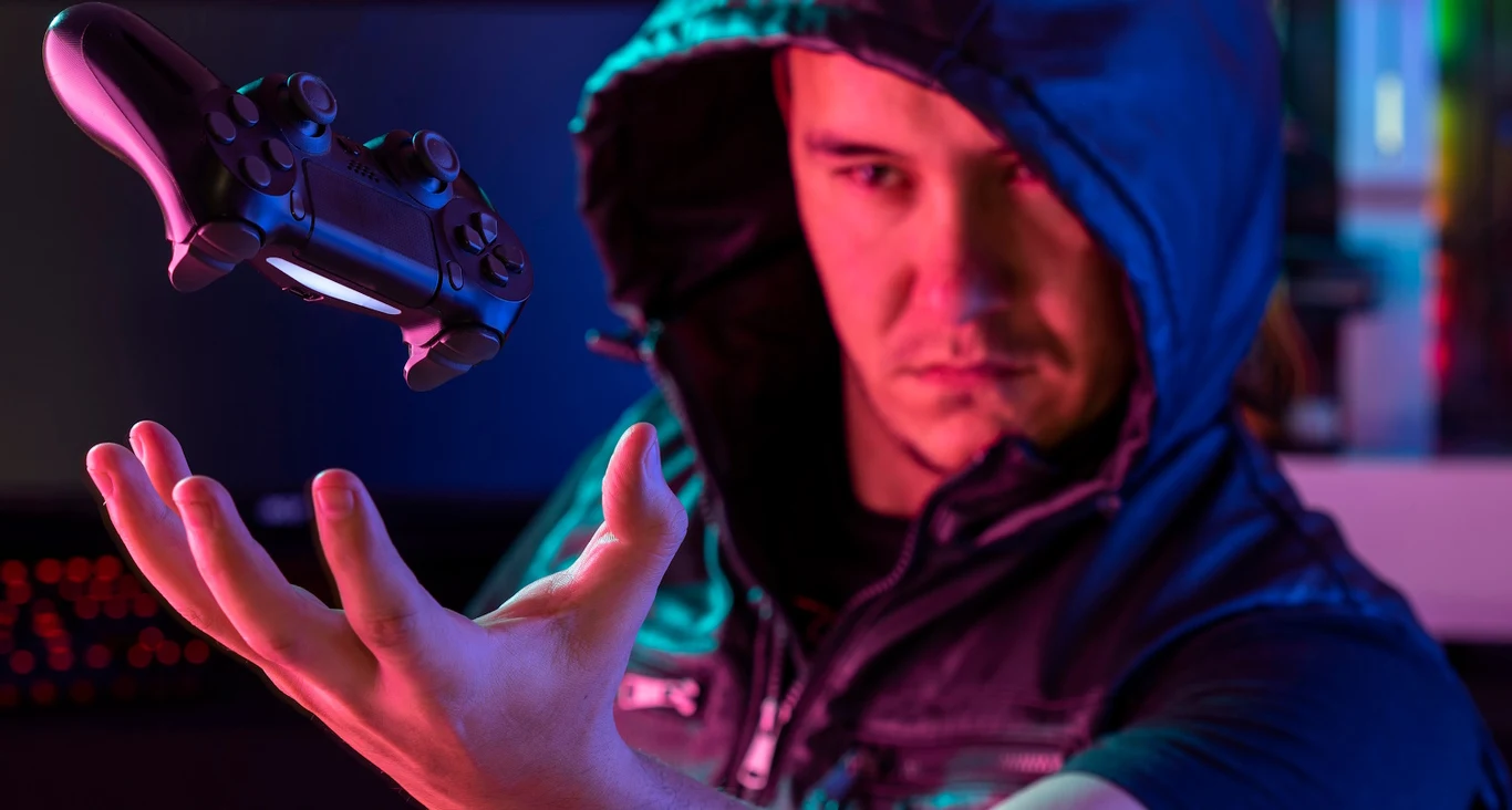 Najciekawsze propozycje RPG z 2025 Mężczyzna w ciemnej bluzie z kapturem unosi rękę ku górze, a nad nią lewituje kontroler do konsoli do gier, całość utrzymana w kolorowych światłach tworzących futurystyczny klimat.