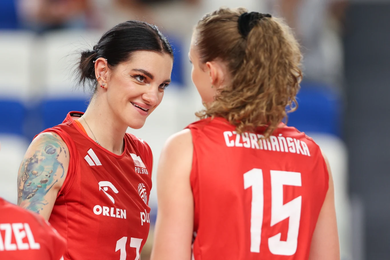 Dwie siatkarki w czerwonych koszulkach drużyny narodowej Polski rozmawiają ze sobą na boisku. Jedna z zawodniczek ma widoczne tatuaże na ramieniu, obie ubrane w stroje sportowe z numerami i nazwiskami, w tle widownia lekko rozmyta.