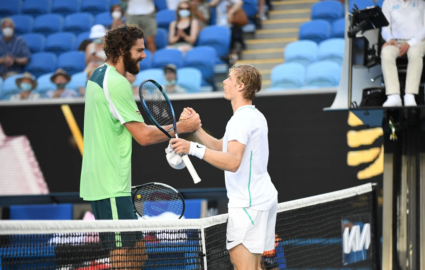 Dwóch tenisistów gratuluje sobie po zakończonym meczu przy siatce na korcie, w tle widoczne niebieskie trybuny z publicznością.
