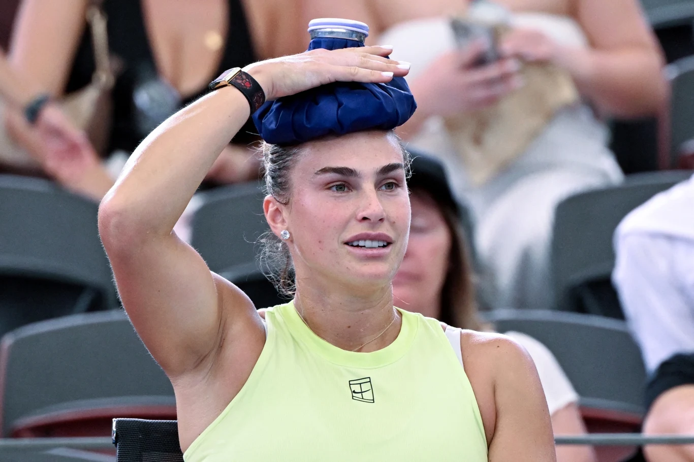 Aryna Sabalenka Kobieta w sportowym stroju siedzi na trybunach i trzyma worek z lodem na głowie, wokół niej widoczne są zamazane sylwetki widzów.