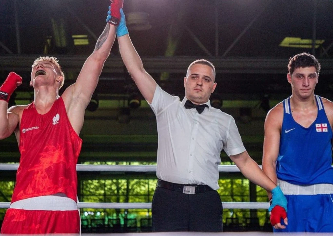 Sędzia unosi rękę zwycięskiego boksera w czerwonym stroju, podczas gdy drugi zawodnik w niebieskim stroju stoi z boku ze smutnym wyrazem twarzy.