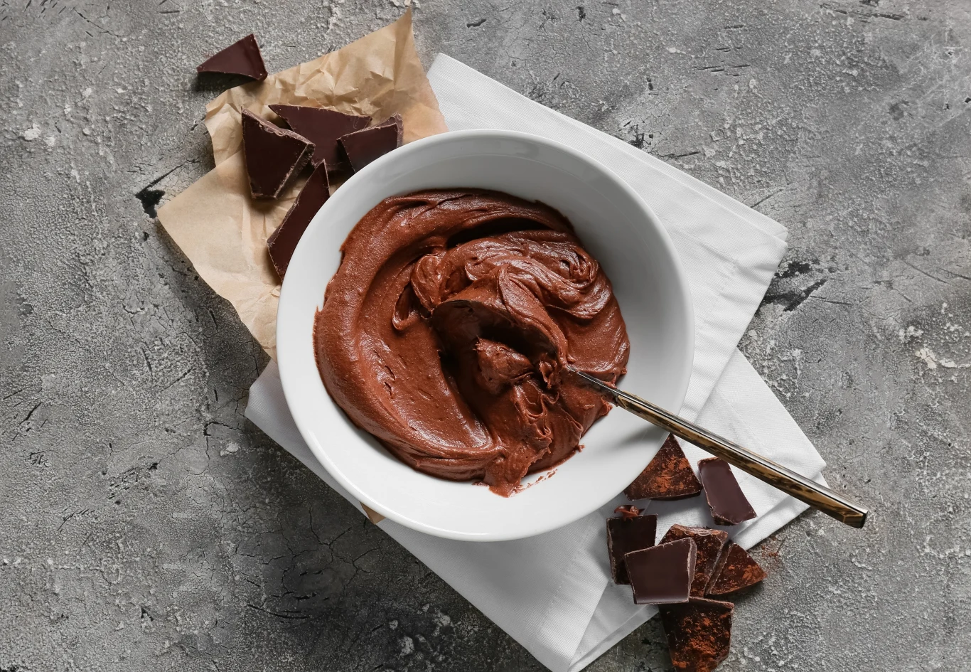 Smakuje i wygląda jak klasyczny mus czekoladowy, a jest zdrowszy i pełen białka Miseczka z gęstym kremem czekoladowym i łyżką, otoczona kawałkami gorzkiej czekolady na szarej powierzchni. Część czekolady leży na papierze, obok skorup czekoladowych. Dominują odcienie brązu i szarości.