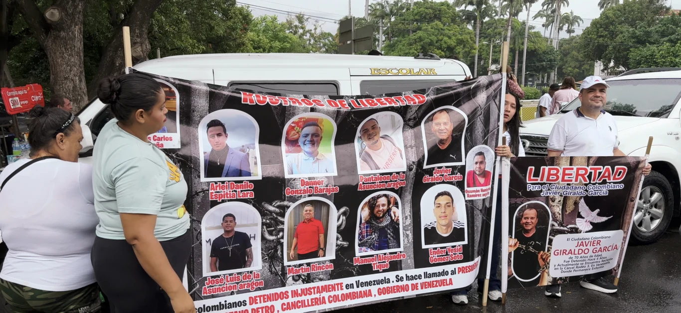 Kilka osób trzyma duży transparent ze zdjęciami i nazwiskami kilku osób oraz wezwaniem do uwolnienia więźniów. Transparent zawiera napisy w języku hiszpańskim oraz zdjęcia ludzi, którzy prawdopodobnie są więzieni lub zaginieni. W tle znajduje się samoc...