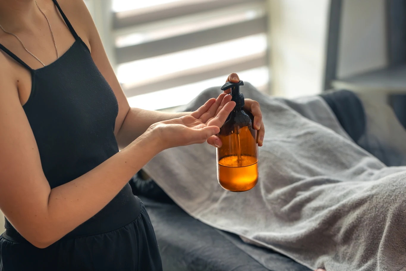 Kobieta w ciemnej koszulce nalewa olejek z brązowej butelki na dłoń, w tle leży przykryta ręcznikiem osoba na stole do masażu.