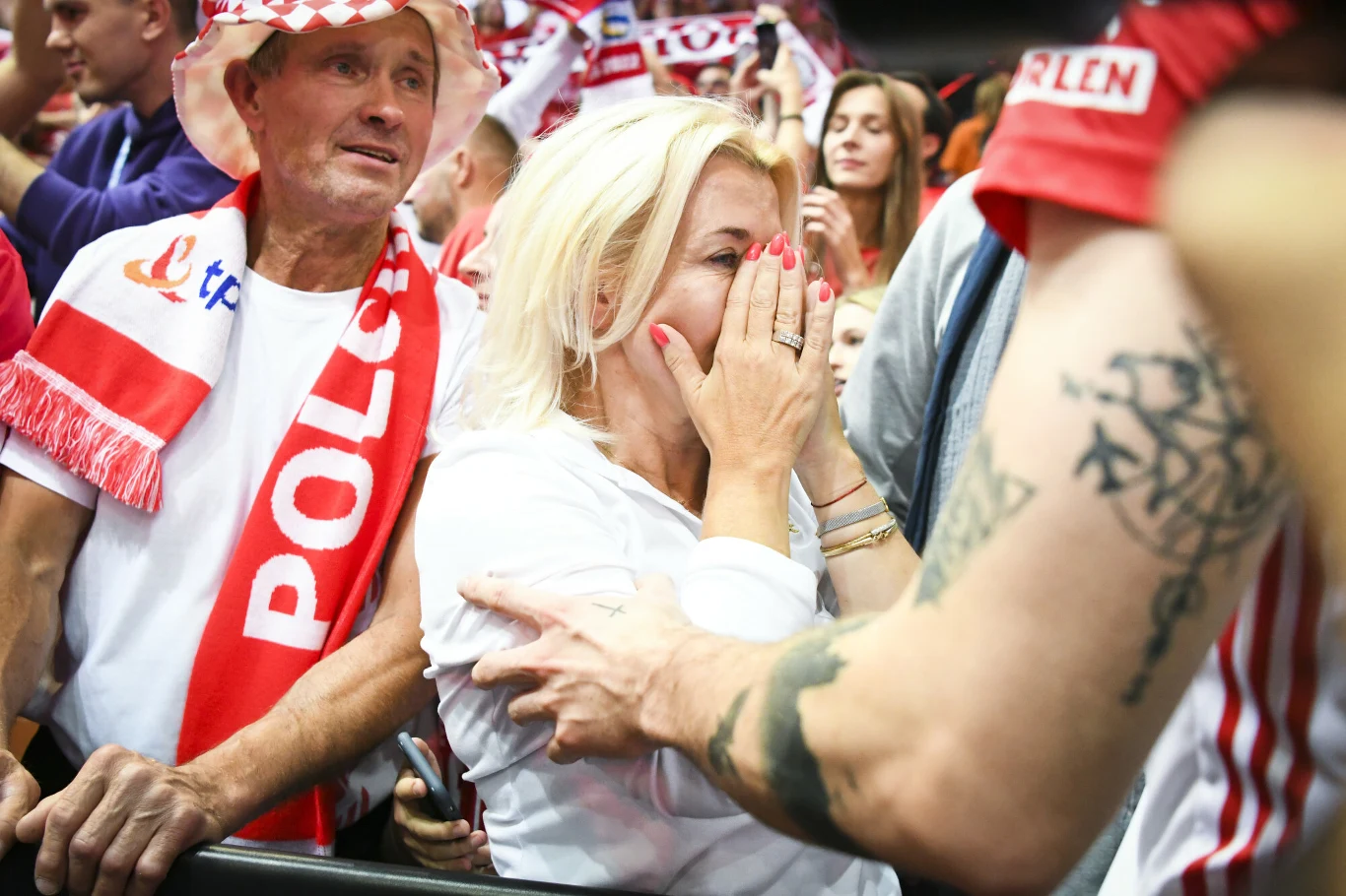 Kibice w biało-czerwonych strojach dopingują swoją drużynę, kobieta o blond włosach zakrywa usta dłońmi z wyrazem silnych emocji, wokół niej inni fani entuzjastycznie uczestniczą w wydarzeniu sportowym.