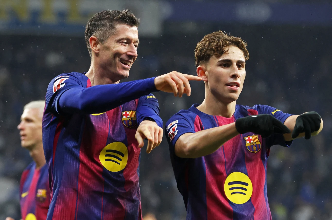 Dwóch piłkarzy FC Barcelony ubranych w granatowo-bordowe stroje z żółtymi logotypami cieszy się po zdobyciu gola, wskazując palcami w stronę trybun. W tle widać rozmazany stadion oraz kibiców.