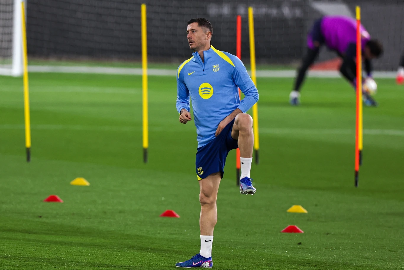 Piłkarz ubrany w niebieski dres trenuje na boisku otoczonym kolorowymi pachołkami i tyczkami, w tle widoczna sylwetka bramkarza podczas rozgrzewki.