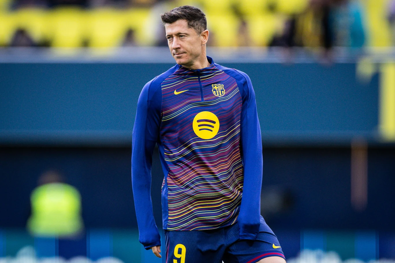 Piłkarz ubrany w sportowy strój klubu FC Barcelona z numerem 9 przygotowuje się do meczu na tle stadionu z żółto-niebieskimi trybunami.