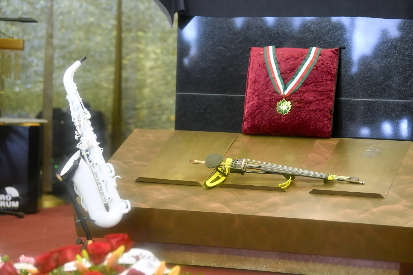Biały saksofon ustawiony na stojaku obok instrumentu smyczkowego oraz medalu na czerwonej poduszce. W tle elegancka dekoracja i elementy sceniczne. Na pierwszym planie widoczne są fragmenty czerwonych róż.