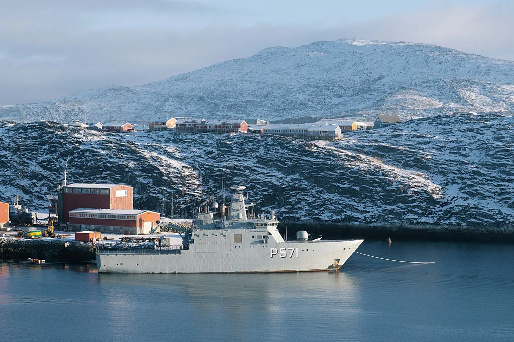 Okręt wojskowy zacumowany przy nabrzeżu na tle ośnieżonych wzgórz i kilku budynków w małym miasteczku. Woda spokojna, a klimat chłodny i zimowy. Nad okolicą widać rozproszone kolorowe domy oraz większy czerwony budynek industrialny.