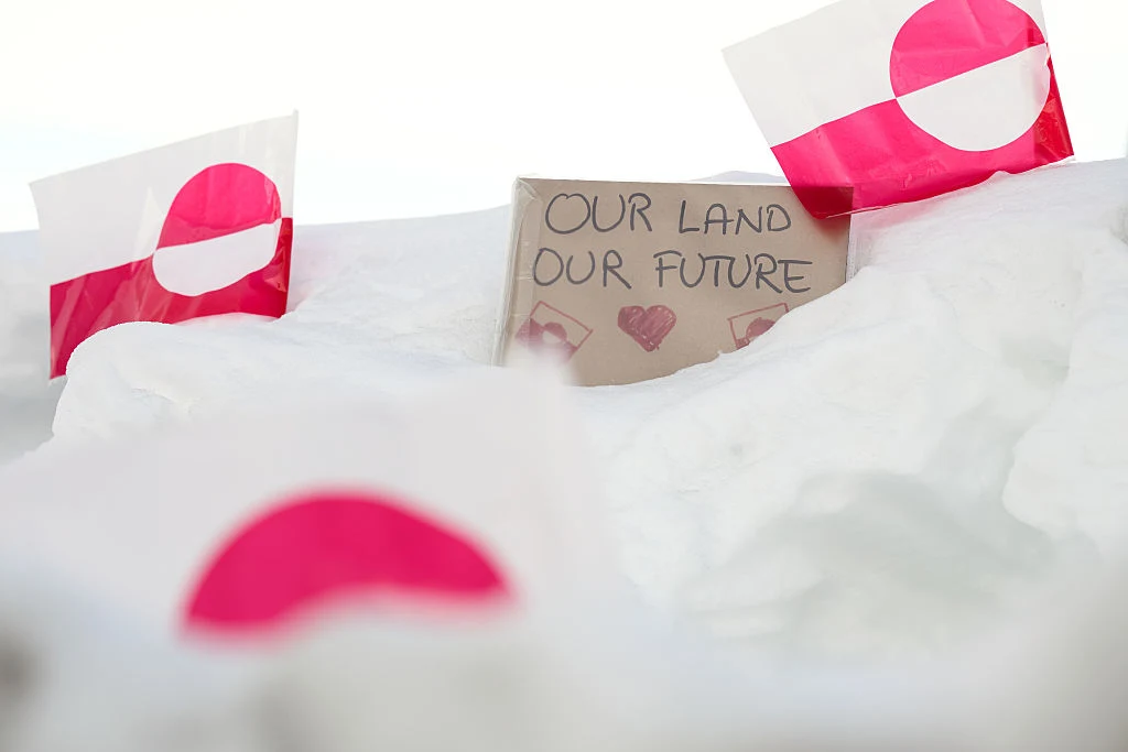 Cztery flagi Grenlandii wbite w śnieg oraz kartka z napisem 'Our land Our future' i narysowanym sercem, podkreślająca znaczenie ziemi i przyszłości dla mieszkańców.