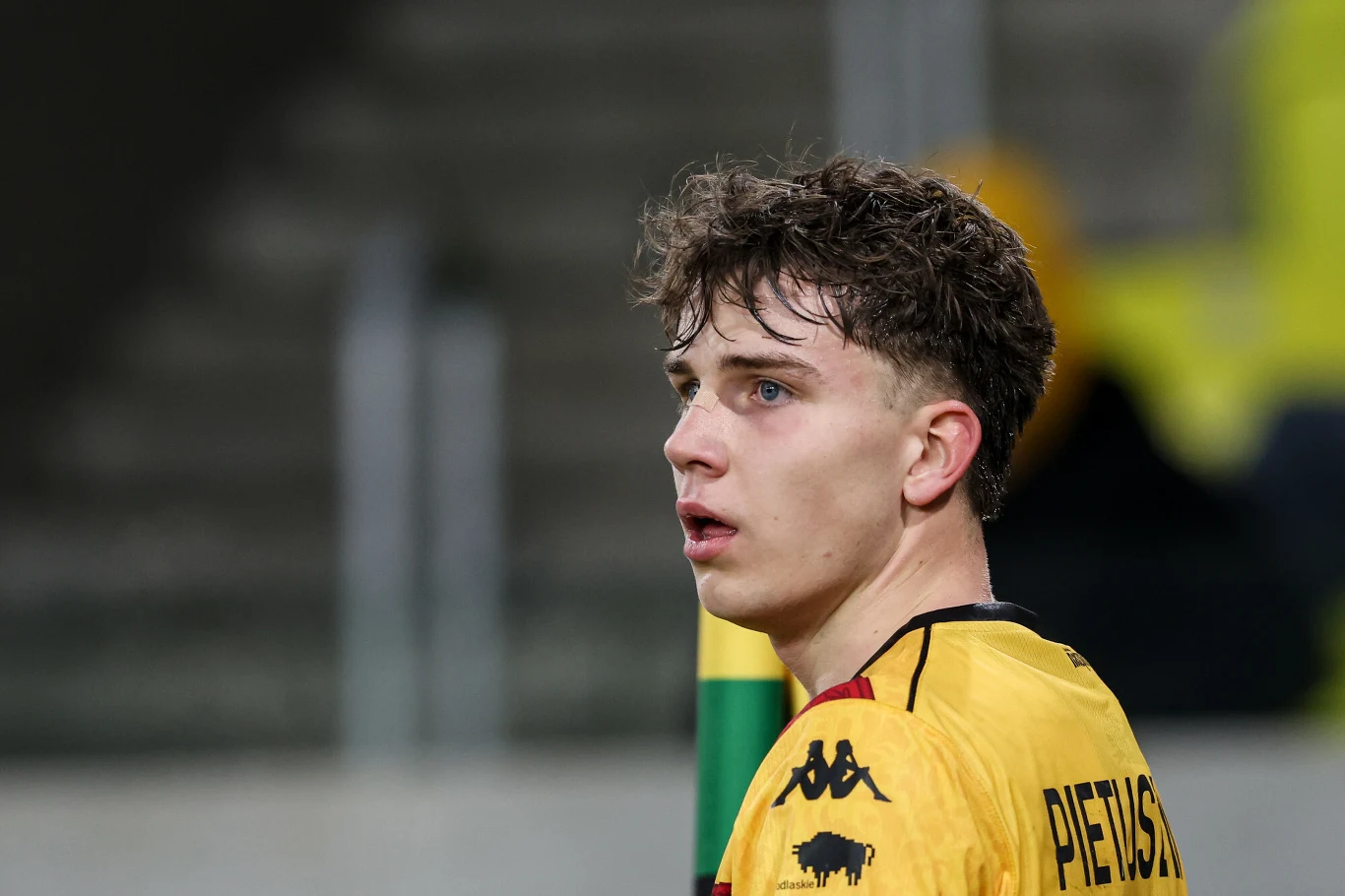 Młody mężczyzna w żółtej koszulce sportowej z nazwiskiem 'Pietrzyk' na plecach patrzy w bok, w tle rozmyte schody stadionu i osoba w żółtej kurtce.