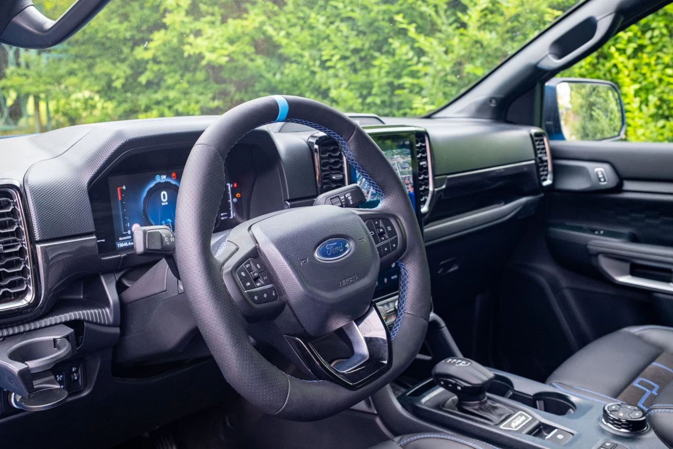 Kokpit nowoczesnego samochodu marki Ford z cyfrowym wyświetlaczem na desce rozdzielczej, skórzaną kierownicą z niebieskim akcentem oraz zaawansowanym panelem sterującym, w tle widoczne siedzenie pasażera i drzwi.