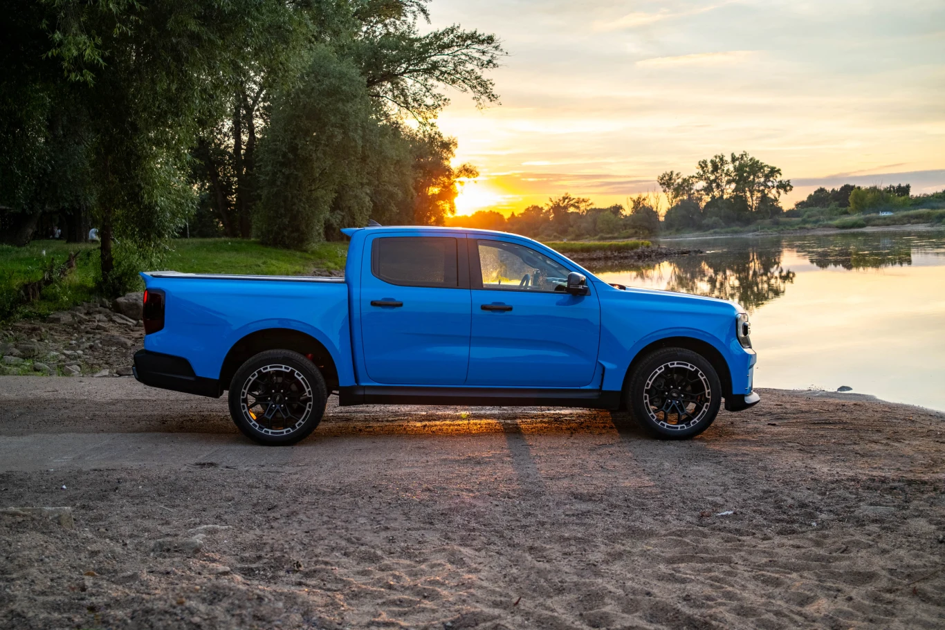 Niebieski pick-up zaparkowany na piaszczystej plaży nad rzeką o zachodzie słońca, w tle drzewa oraz spokojna tafla wody odbijająca promienie słońca.