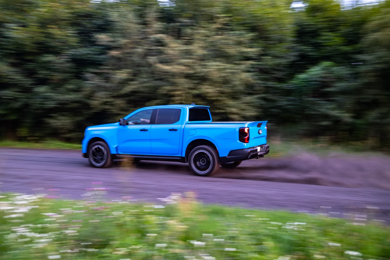 Jasnoniebieski pickup jedzie z dużą prędkością po nieutwardzonej drodze, pozostawiając za sobą chmurę czarnego dymu, w tle widać drzewa i rozmyte zielone krzewy.