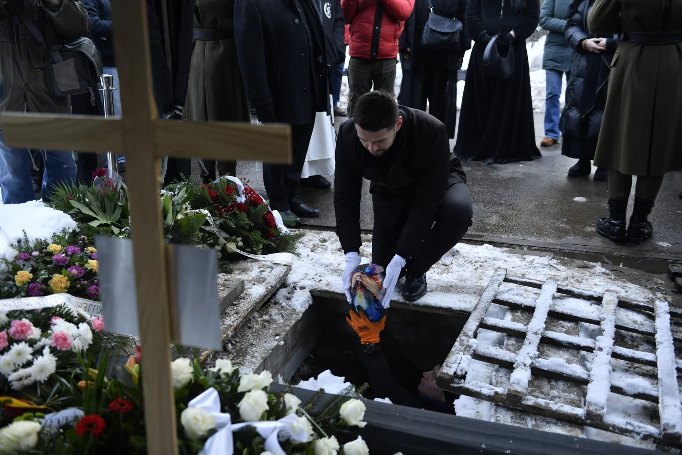 Człowiek pochylony nad grobem przekazuje przykrytą flagą rzecz osobie znajdującej się w dole, wokół grób udekorowany kwiatami, z drewnianym krzyżem i grupą ludzi stojących w tle, zimowa sceneria ze śniegiem na ziemi.