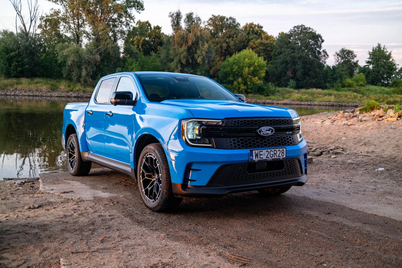 Niebieski samochód typu pick-up marki Ford stoi zaparkowany na piaszczystej nawierzchni nad brzegiem rzeki, w tle widoczna zieleń drzew oraz spokojna tafla wody.