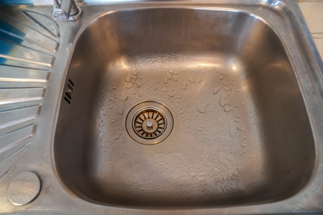 Problemy z odpływem pojawiają się w wielu domach. Sprawdź, jak sobie z tym poradzić Stalowy zlew kuchenny z widocznymi kroplami wody i odpływem pośrodku, otoczony metalowym blatem.