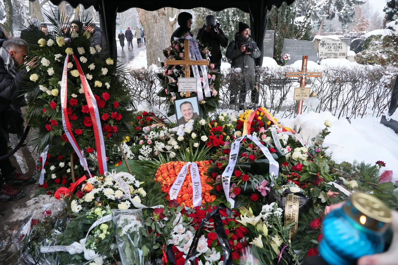 Świeże kwiaty oraz wieńce ułożone na grobie otoczonym śniegiem, centralnie położony portret zmarłego, krzyże nagrobne, w tle widoczne osoby i fotografowie, dominujące kolory to biel śniegu oraz czerwień i zieleń kwiatów.