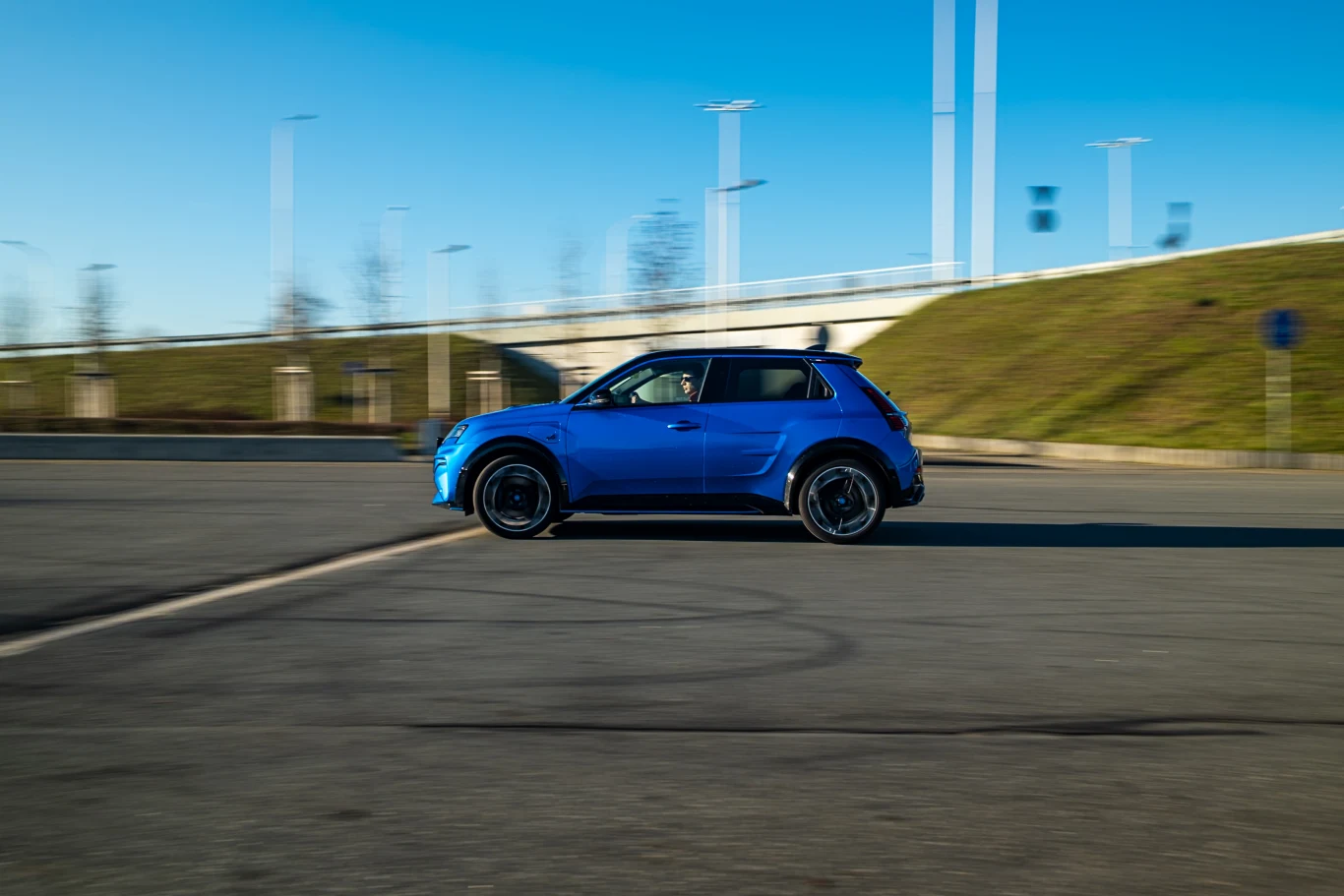 Niebieski samochód osobowy poruszający się po pustym parkingu asfaltowym z widocznym rozmyciem tła wskazującym na ruch. W tle nasyp ziemny, kilka lamp ulicznych oraz jasne, bezchmurne niebo.