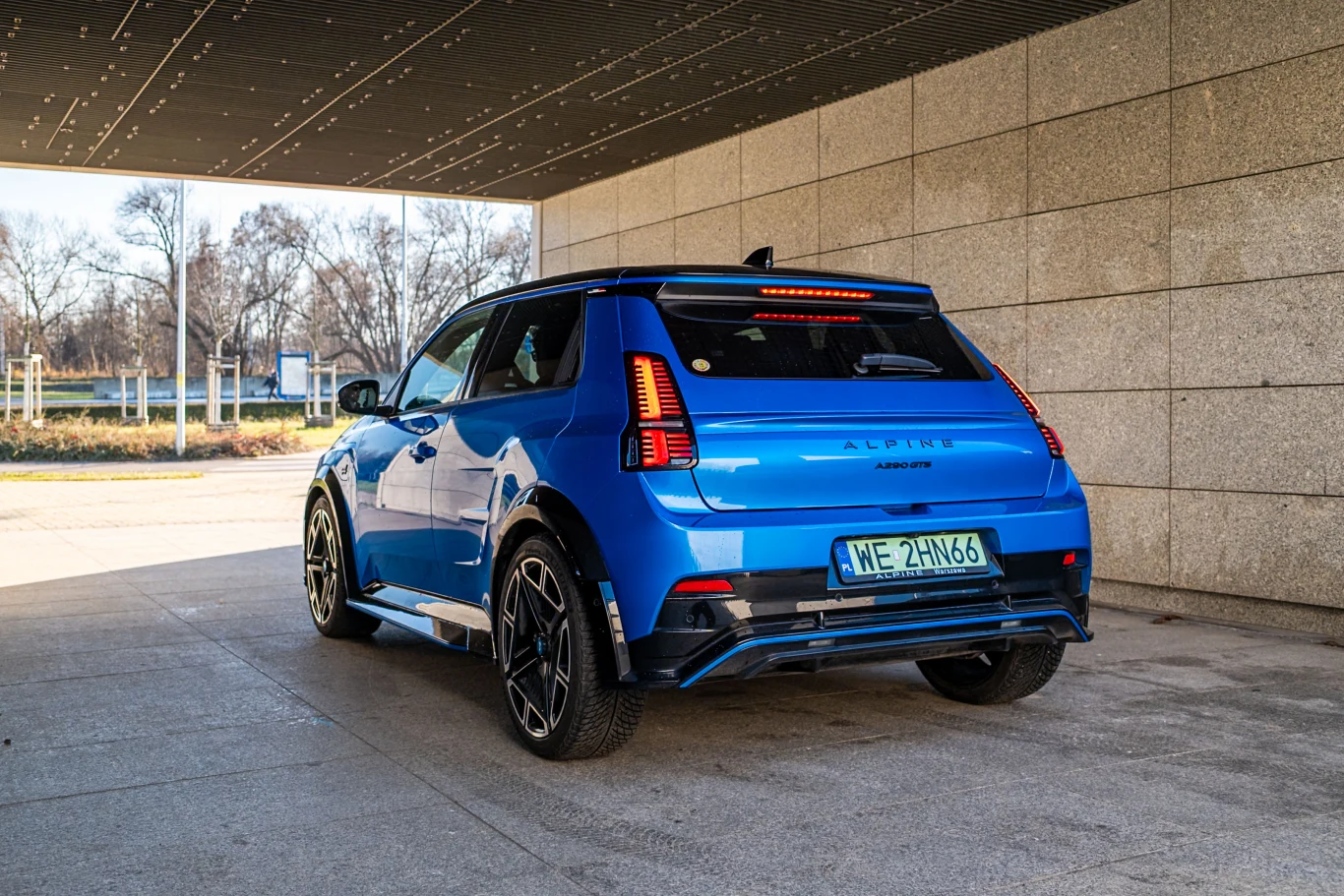 Nowoczesny, niebieski samochód osobowy zaparkowany pod betonowym zadaszeniem, widok z tyłu. Pojazd wyróżnia się sportowym wyglądem, solidnymi felgami i dynamicznymi liniami nadwozia. W tle widoczne są drzewa i fragmenty miejskiej architektury.