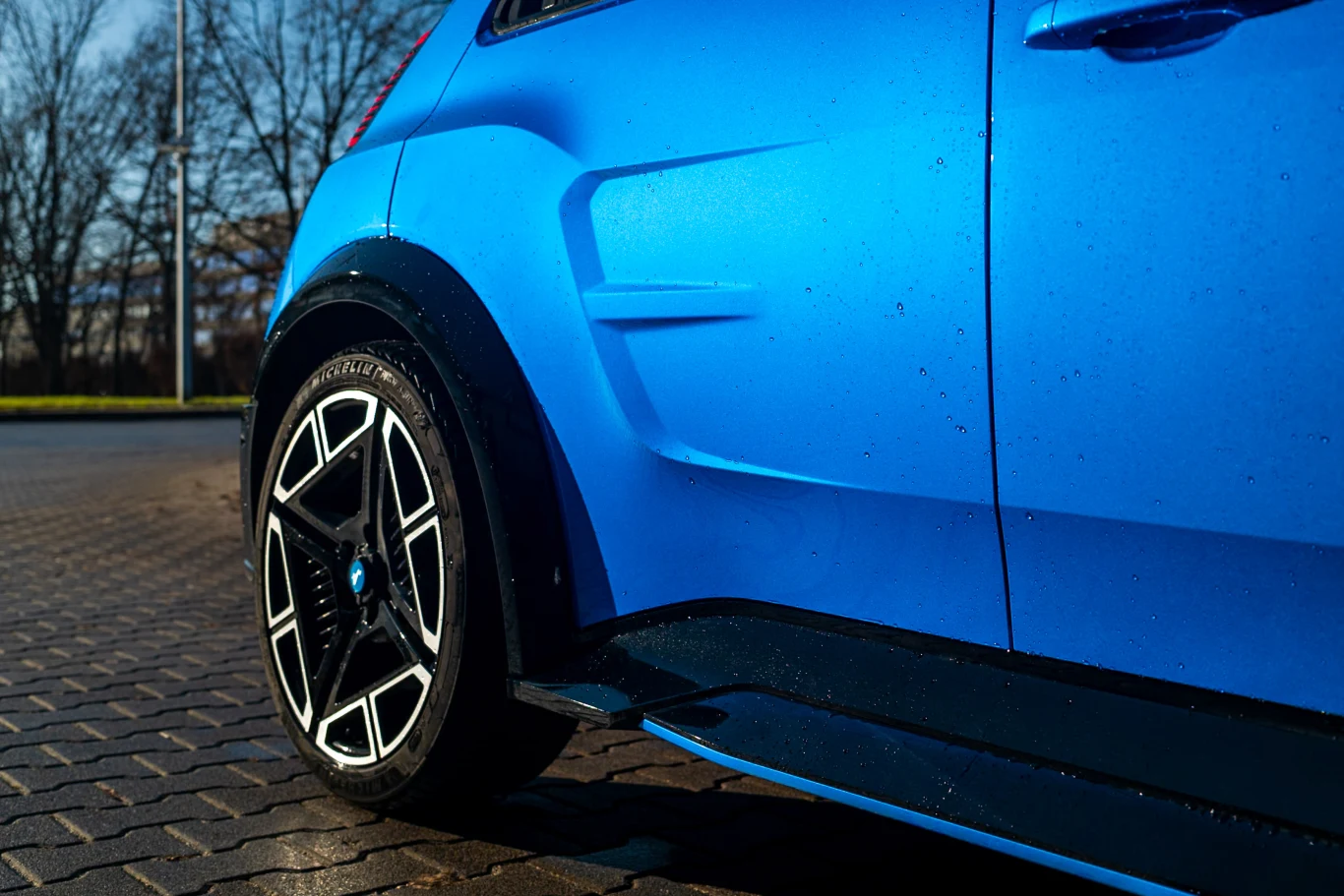 Fragment nowoczesnego, niebieskiego samochodu z widocznym tylnym kołem oraz elementami karoserii, zaparkowanego na kostce brukowej przy ulicy, w tle drzewa bez liści.