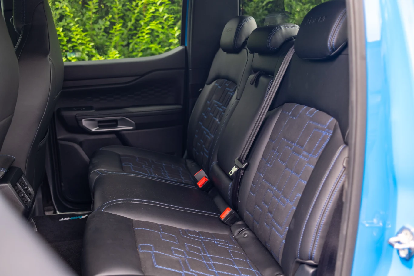 Tylny rząd siedzeń w samochodzie z czarną tapicerką ozdobioną niebieskimi przeszyciami, widocznymi zagłówkami oraz pasami bezpieczeństwa, po lewej stronie widoczne drzwi z uchwytem.