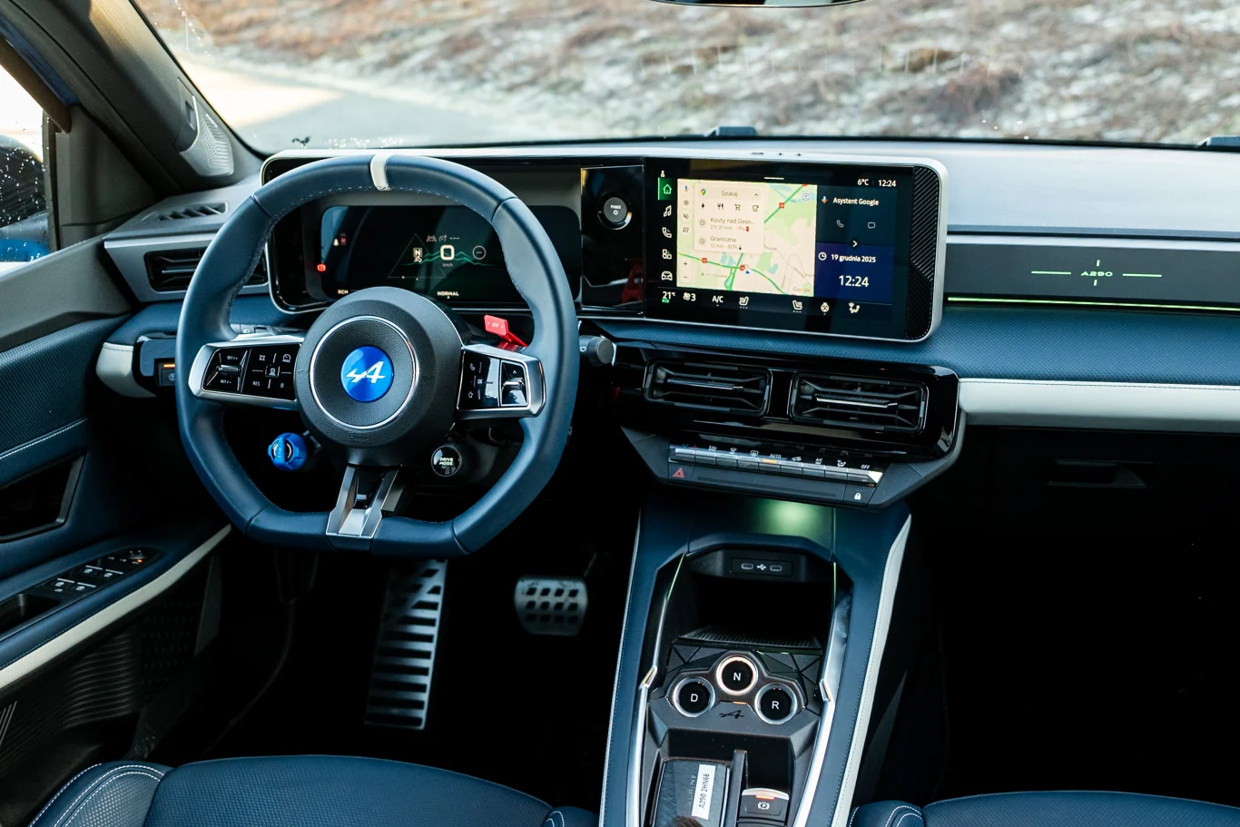 Wnętrze nowoczesnego samochodu Alpine z wyraźnie widoczną kierownicą, cyfrowym kokpitem oraz dużym ekranem systemu multimedialnego z nawigacją. Układ przycisków na środkowej konsoli i sportowy design podkreślają nowoczesność pojazdu.