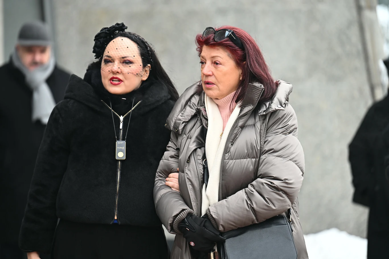 Dwie kobiety ubrane w zimowe kurtki stoją obok siebie na zewnątrz, jedna z nich ma na głowie ozdobę oraz widoczną biżuterię, trzymają się pod ramię, w tle widać rozmazane postacie i fragment betonowej ściany.