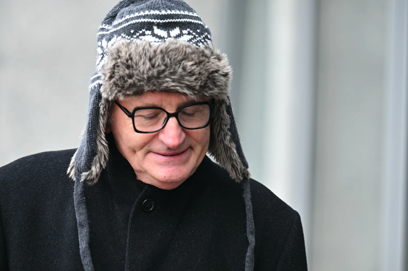 Uśmiechnięty mężczyzna w okularach, ubrany w ciemny płaszcz i zimową czapkę z futrzanym obszyciem, stoi na tle jasnej powierzchni.