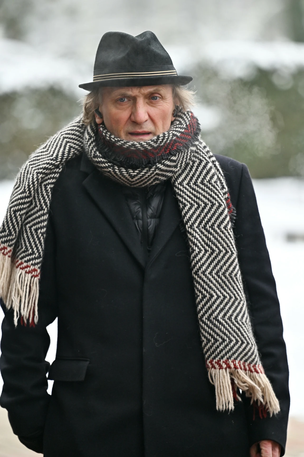 Mężczyzna w ciemnym płaszczu i kapeluszu, z dużym wełnianym szalikiem w biało-czarne wzory z frędzlami, stoi zimą na tle zaśnieżonego krajobrazu.