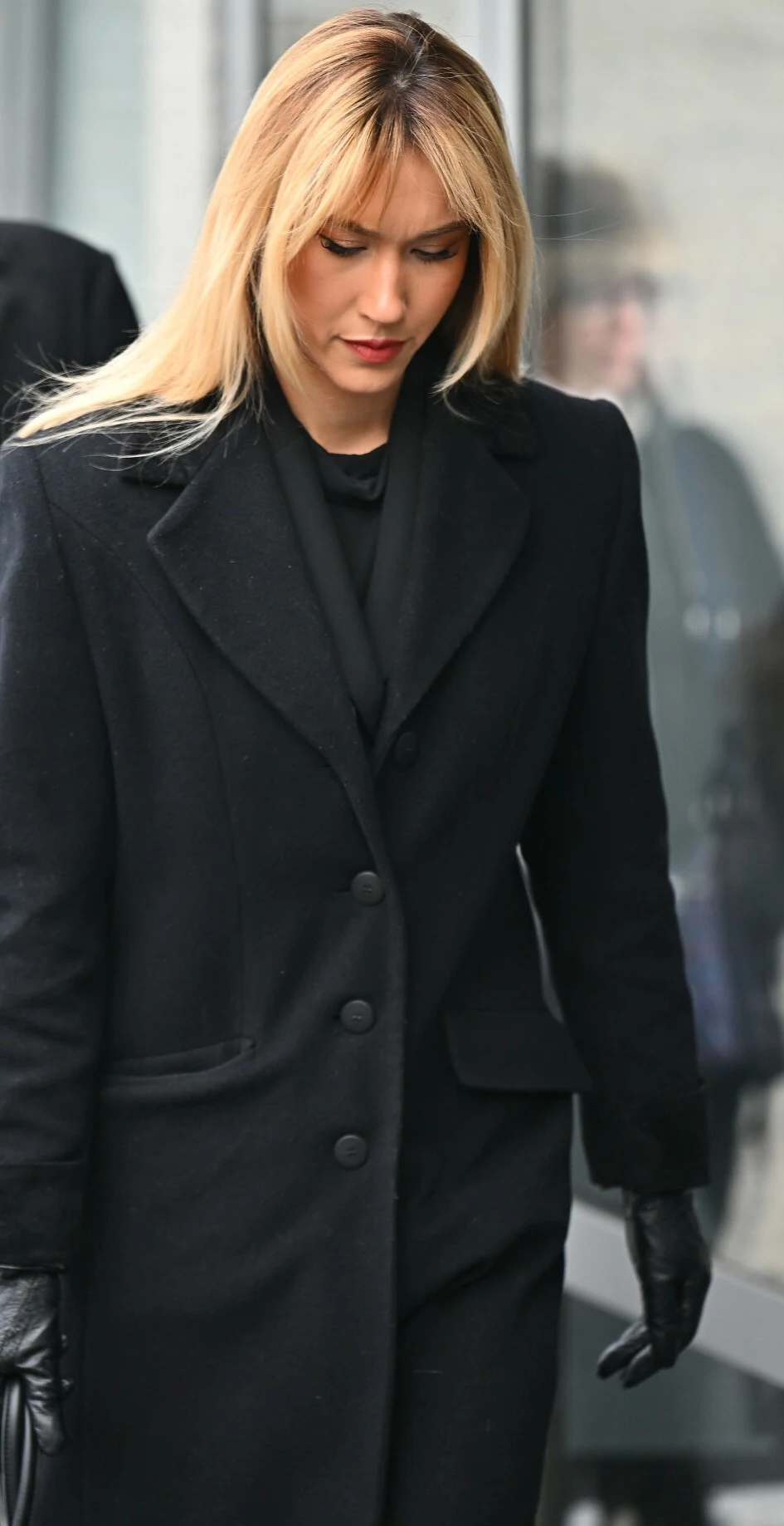 Kobieta o blond włosach ubrana w elegancki czarny płaszcz i czarne rękawiczki, stoi w pobliżu przeszklenia, spoglądając w dół.