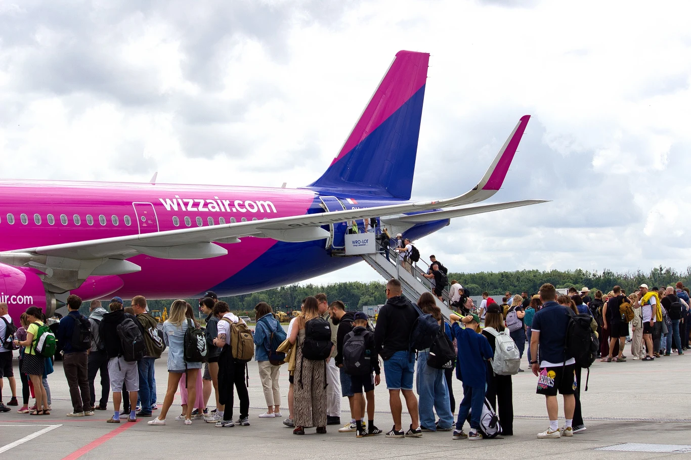 Pasażerowie ustawieni w kolejce na płycie lotniska wchodzą po schodach do samolotu linii Wizzair o charakterystycznych różowych i fioletowych barwach. W tle widoczny fragment pasa startowego oraz zalesiony horyzont pod pochmurnym niebem.