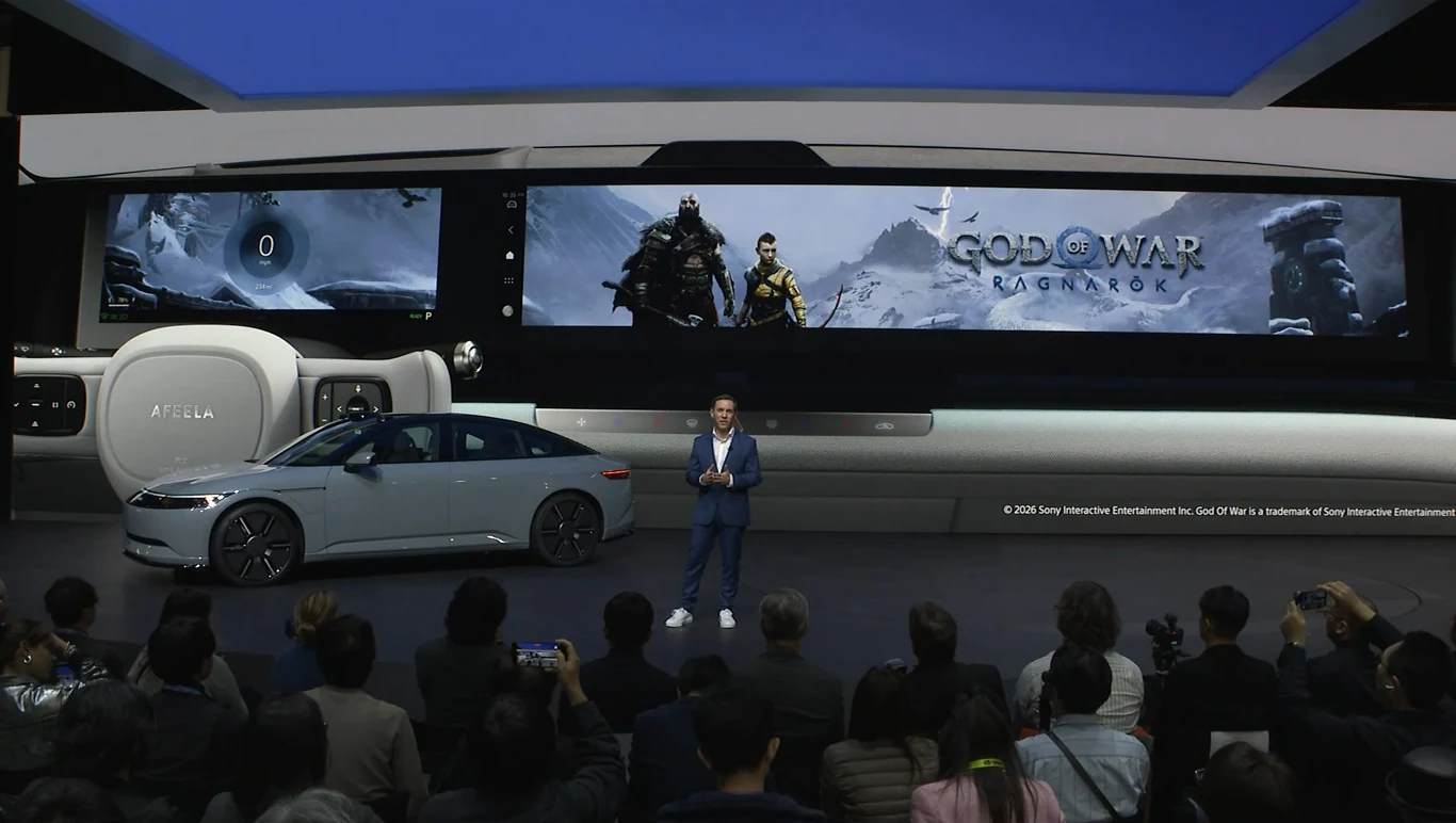 Auto przyozdobione motywami z serii God of War z pewnością przypadnie do gustu fanom marki Mężczyzna w garniturze występuje na scenie przed publicznością, obok stoi nowoczesny samochód, a w tle na dużym ekranie widoczna jest grafika z gry God of War Ragnarok.